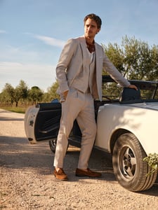 Ein Bräutigam steht in einem Anzug im Boho-Stil mit braunen Mokassins vor einem weißen Cabriolet.