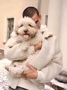 Ein Mann, welcher einen cremefarbenen Hoodie und eine Teddyjacke trägt. Er hält einen Hund in seinen Arm und kuschelt mit ihm.