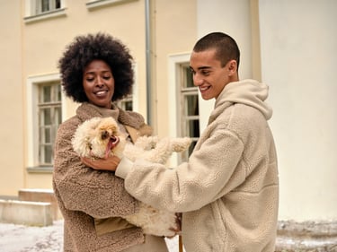 Ein Mann und eine Frau, welche jeweils eine Teddyjacke tragen. Die Frau hält einen Hund in ihren Armen und der Mann streichelt ihn. 