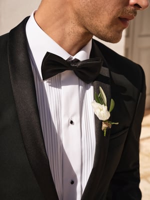A groom in a nice black suit who is wearing a bow tie and a flower brooch.